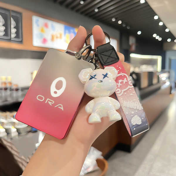 A hand holds the ora iq key cover 2024, a sleek gradient red and silver leather remote sleeve, accompanied by a cute white be...