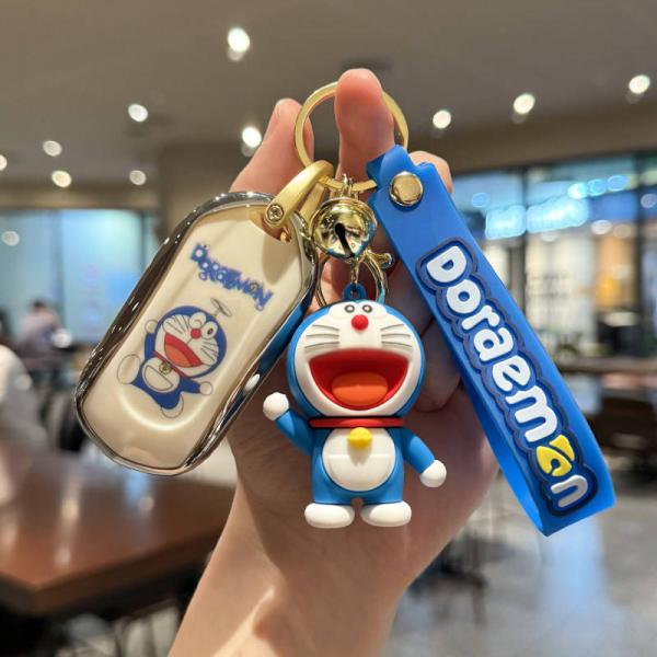 A hand holds a doraemon-themed car key cover and accessories, featuring a white key fob with a doraemon print, a blue doraemo...