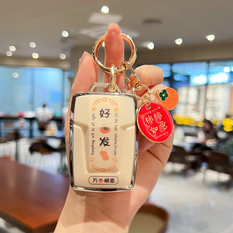 A hand holds a stylish tank 300 car key cover with and text, accompanied by a red charm with design, set in a coz...