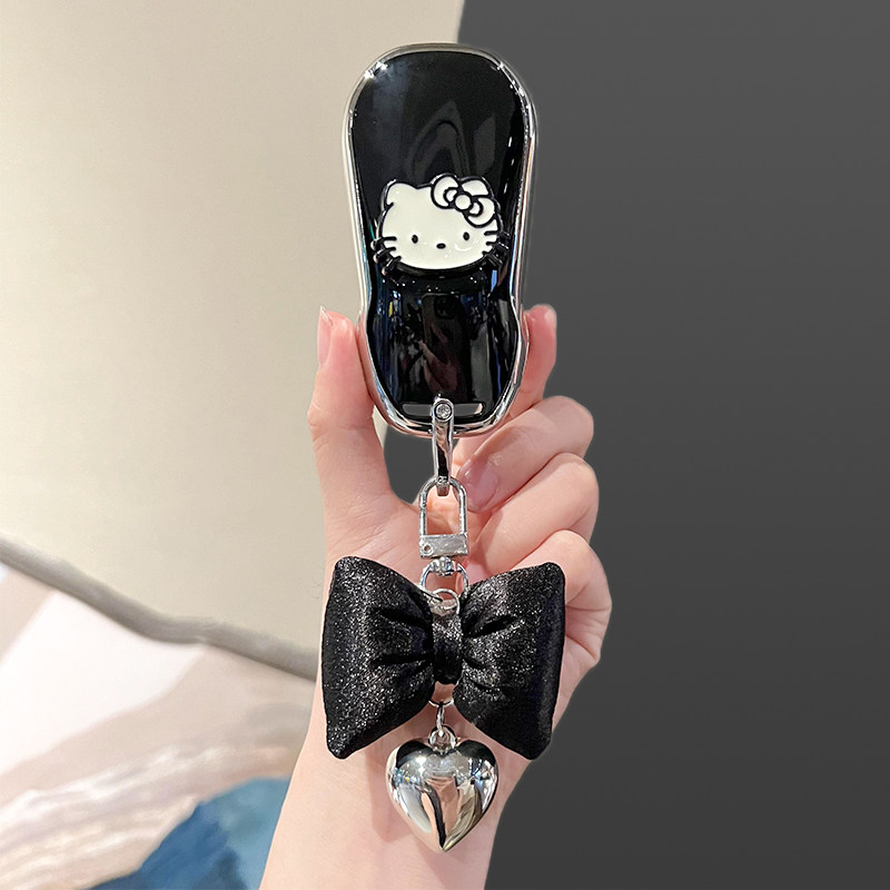 A hand holds a sleek black key cover featuring a white hello kitty design, adorned with a shiny black bow and silver heart ch...