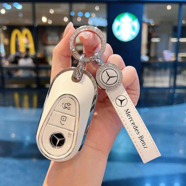 A hand holds a mercedes-benz key cover with a sleek white design, featuring a mercedes logo and a matching keychain, set agai...