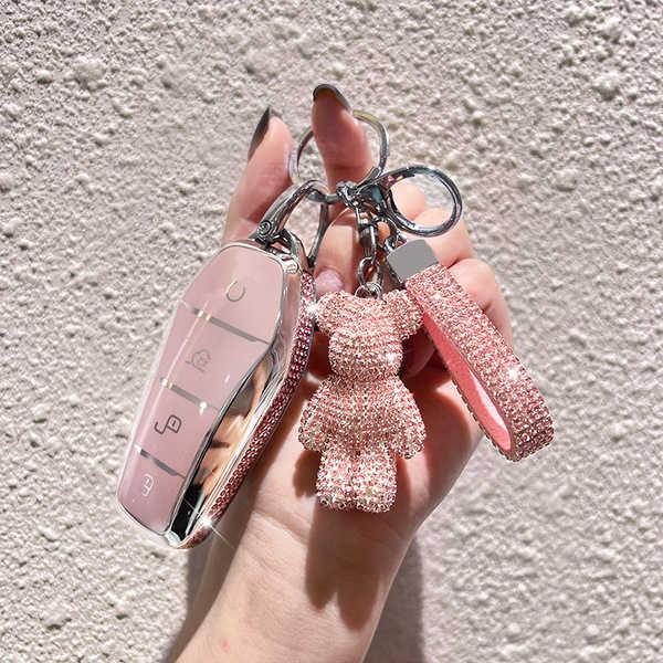 A hand holds a pink byd key cover adorned with rhinestones, featuring a matching teddy bear charm and wrist strap, designed f...