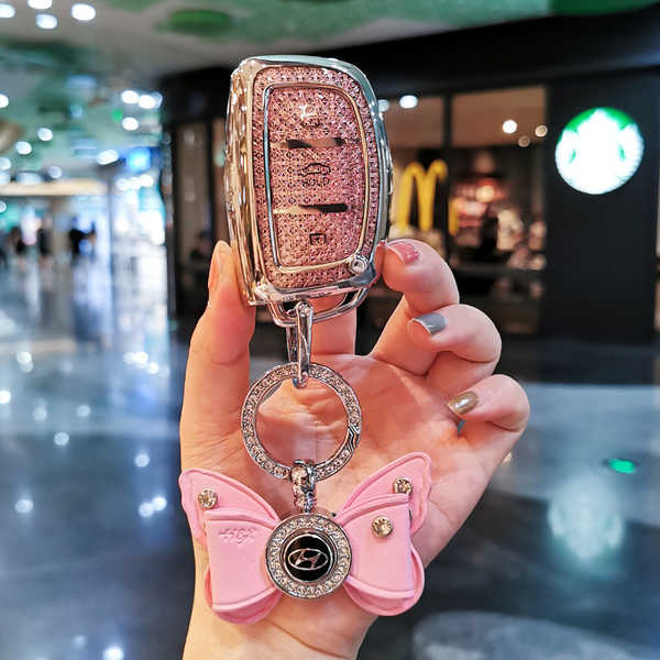 A hand holds a sparkling pink hyundai key cover for ix35, elantra & tucson, adorned with a butterfly charm and a glittery fin...