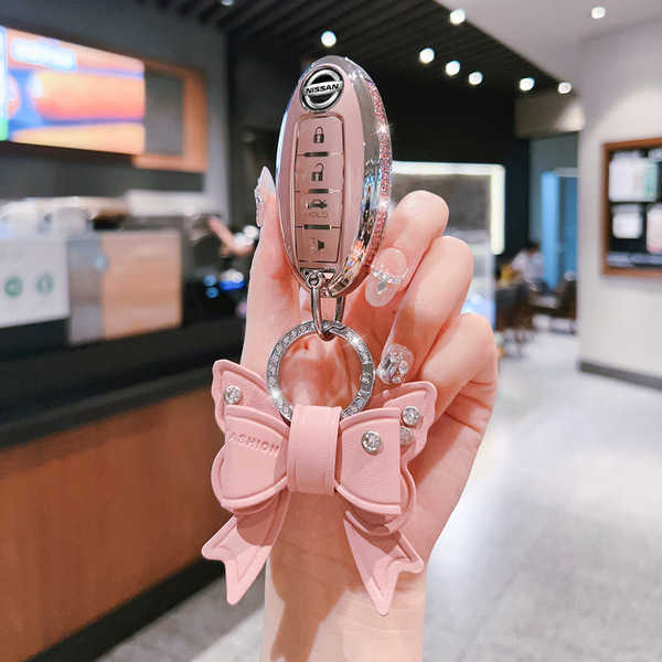 A hand holds a nissan key fob with a pink bow-shaped key cover, featuring decorative rhinestones, in a modern café setting.