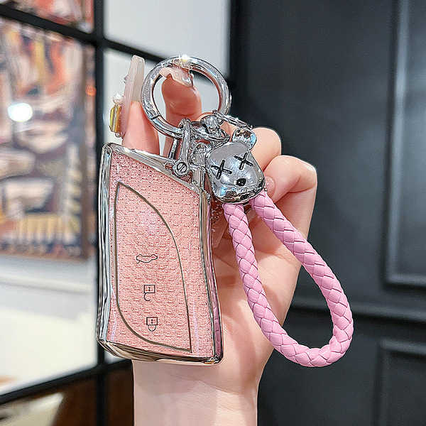 A hand holds a pink lexus key cover with a glittery finish, featuring a silver outline and a braided pink strap with a decora...