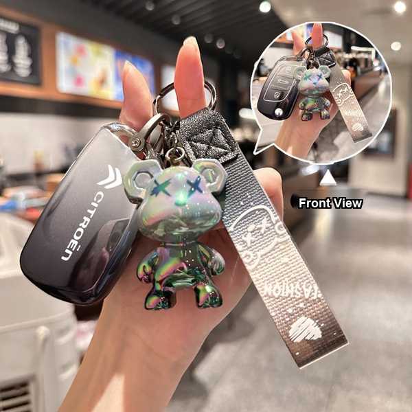 A hand holds a citroën key cover with a shiny, iridescent bear charm and a textured strap, showcasing both the front view and...