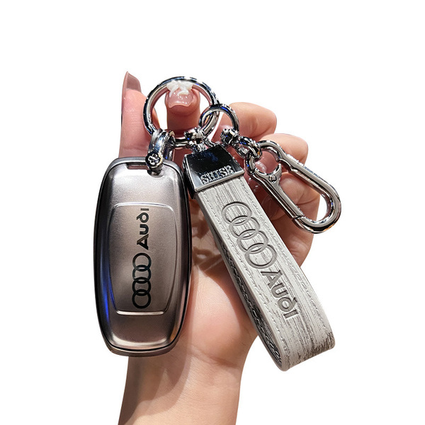 A hand holds a sleek, metallic audi key cover with the iconic four rings logo, paired with a matching leather keychain strap,...