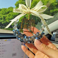 A hand holds a crystal peace car pendant, featuring a gold charm, blue crystal beads, and a white ribbon with love is et...