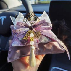 A hand holds a decorative bamboo charcoal car odor remover bag, featuring a purple ribbon with love is everywhere text, a gol...