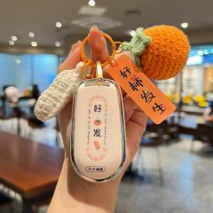 A hand holds a stylish car key cover featuring a good luck charm, a knitted orange fruit, and a corn cob accessory, perfect f...