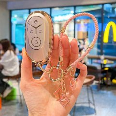 A hand holds a stylish geely galaxy l7 key cover with a gold finish and decorative charms, featuring a pink braided strap, se...