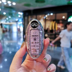 A hand holds a stylish infiniti q50l key cover, featuring a glittery pink design with a sleek silver frame, set against a vib...