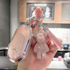 A hand holds a geely key cover with a pink, diamond-encrusted design, featuring a matching teddy bear charm and wrist strap, ...