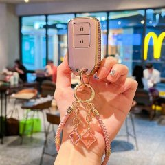 A hand holds a stylish pink honda key cover with gold accents, featuring a lock icon and decorative charms, set against a blu...