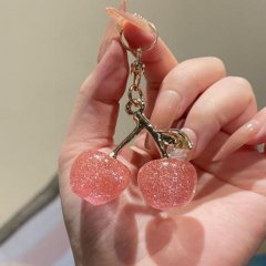 A hand holds a water-drop cherry car air freshener, featuring two glittery pink cherry-shaped pieces with a gold stem and lea...