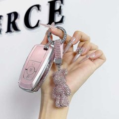A hand holds a pink lincoln corsair key cover, featuring a glittery teddy bear charm and a matching strap, set against a back...