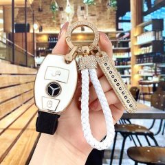 A hand holds a luxurious diamond-embellished mercedes keychain, featuring a white braided rope and a gold-accented design, pe...