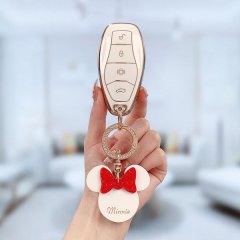 A hand holds a sleek, gold-accented key fob with a minnie mouse charm, featuring a red bow and the name minnie on a white bac...