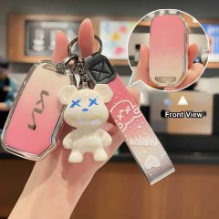 A hand holds a pink kia key cover with a gradient design, featuring a cute bear charm and a patterned strap, shown from the f...