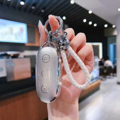 A hand holds a sleek, silver key cover for the ideal l9, featuring a braided white strap and decorative charms, designed for ...