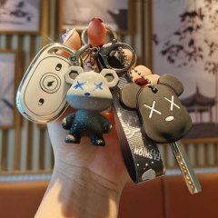 A hand holds a set of key accessories, including a white remote with a circular button and two bear-shaped keychains-one gray...