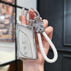 A hand holds a sleek, silver lexus key cover with a textured finish, featuring a white braided lanyard and a decorative keych...