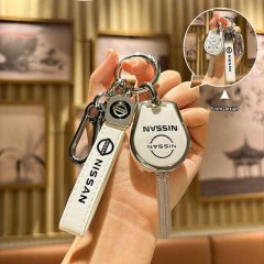 A hand holds a nissan key cover set, featuring a white leather strap and a metallic key fob with the nissan logo, designed fo...