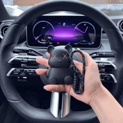 A hand holds a black cute little ink bear key fob cover, featuring x eyes and a rope strap, displayed in front of a cars stee...