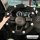 A hand holds a black cotton linen steering wheel cover adorned with white daisy patterns, designed for non-slip grip on an au...
