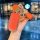 A hand holds a vibrant orange aito m9 key cover with a textured finish, gold accents, and decorative charms, including a meet...