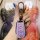 A hand holds a sleek, lavender dongfeng fengguang 580 key cover with a metallic carabiner, set against a vibrant kaws art bac...
