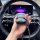 A hand holds a black, bear-shaped silicone key fob cover with a rope strap, displayed in front of a cars steering wheel and d...